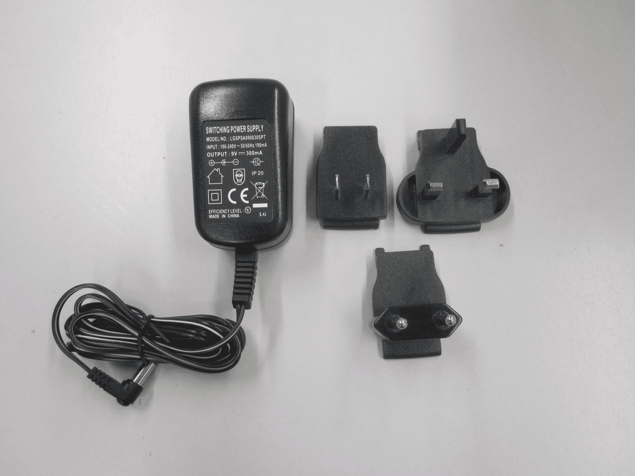 Schwarzes Netzadapter Set auf weißem Hintergrund. Das Set besteht aus einem Stecker mit gefaltetem Kabel und drei Adapter. Die Adapter sind für die Bereiche EU, US und UK.