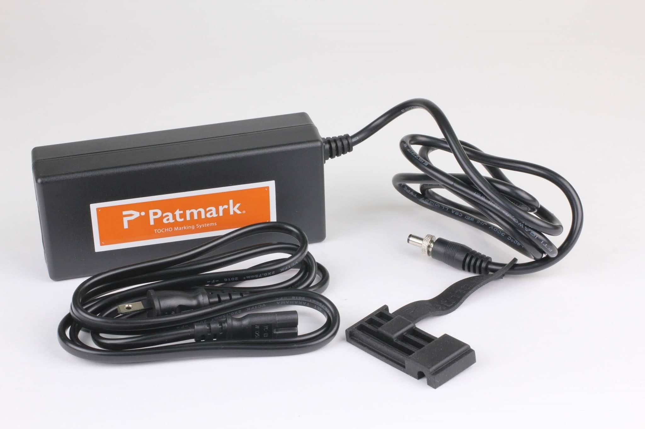 Schwarzer Adapter auf weißem Hintergrund. Der Adapter besteht aus einem schwarzem Transformator was das Logo der Firma Patmark aufweist  und einem Kabel. Vor dem Gerät liegt ein zweites aufgerolltes Kabel.